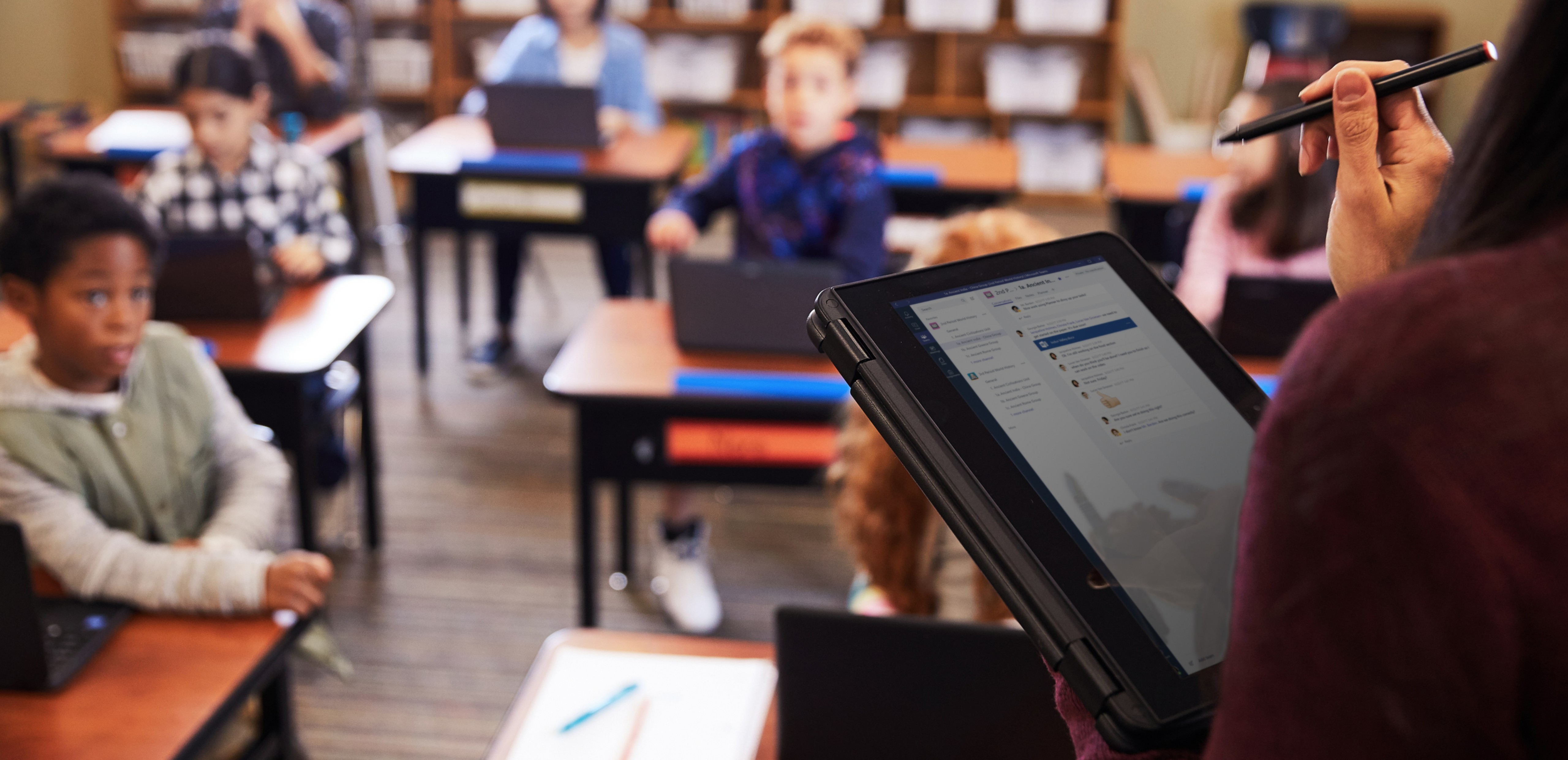 Teacher holding a tablet in a classroom filled with students.