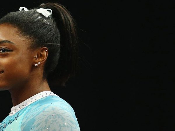 Simone Biles, standing and smiling, facing left, wearing a teal leotard, which is the color of sexual assault awareness.