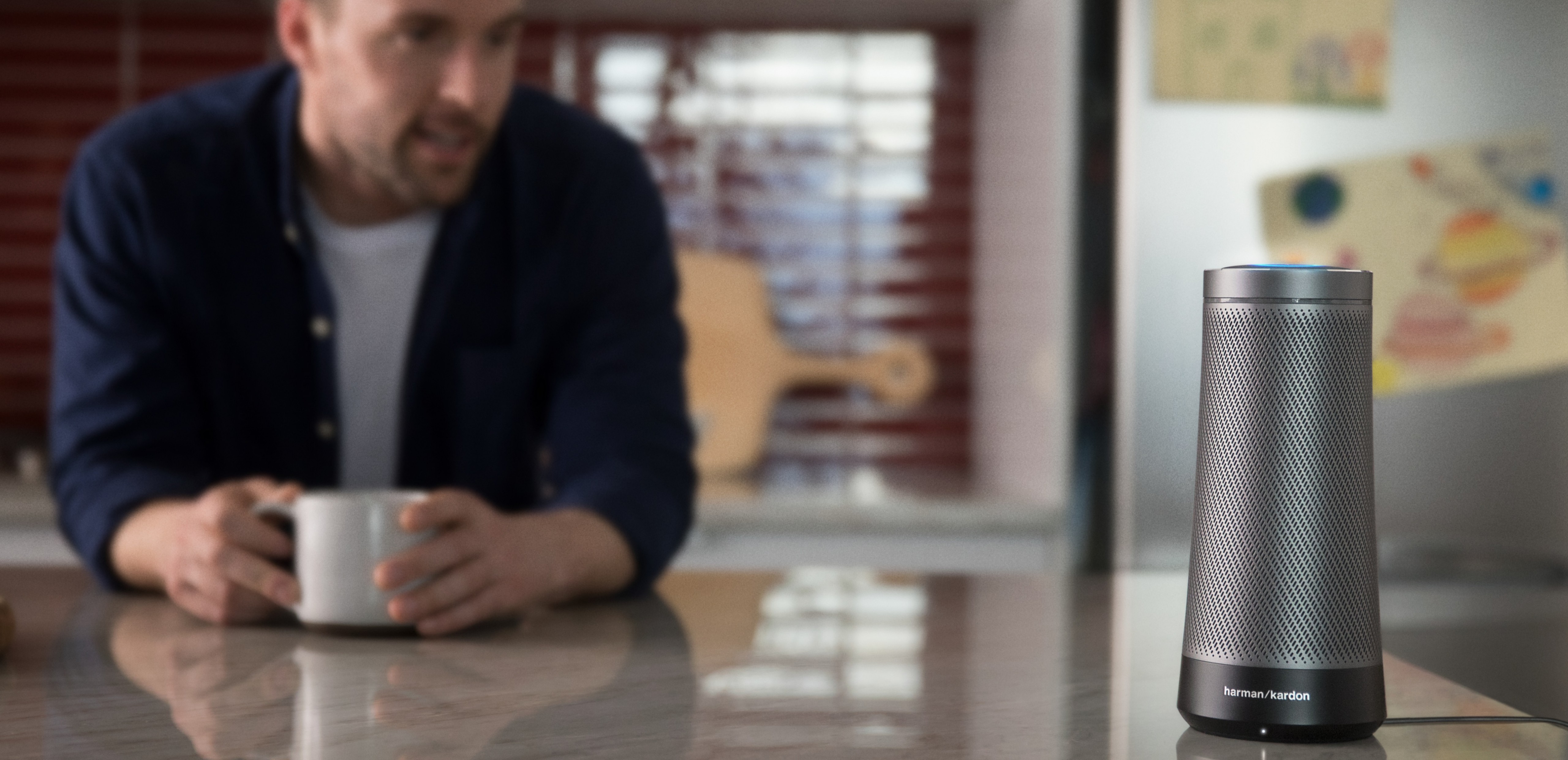 Man standing in kitchen, holding a cup of coffee looking at the Harman Kardon Invoke speaker that combines the rich, captivating sound that Harman Kardon is known for with your personal digital assistant, Cortana, in an innovative and beautifully designed speaker for the home.