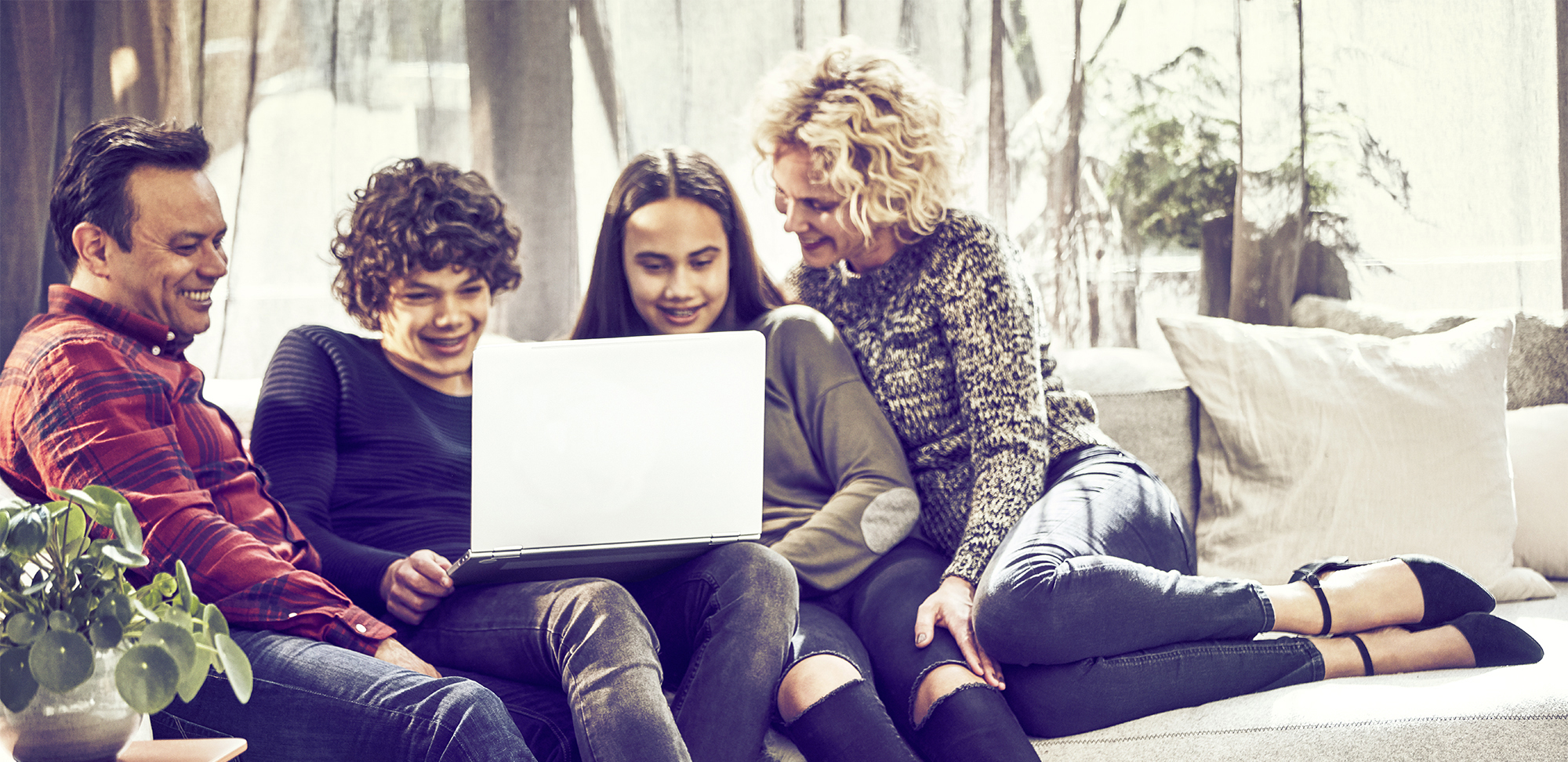 Family of four -- mother, husband, teen son and daughter -- sitting on a couch looking at a silver laptop