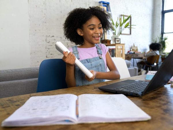 Girl sitting at a table at home laughing at a laptop, textbook in front of her too