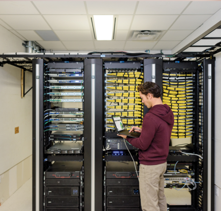 Male IT employee standing in front of server and using Dell laptop. Laptop screen visible with green data analytics chart and graph.