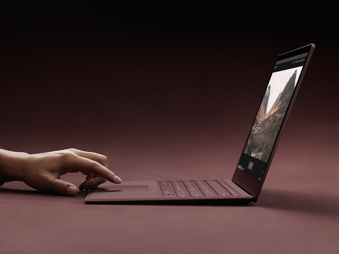 Microsoft Surface Laptop shown in side view in Burgundy color with hand touching the trackpad