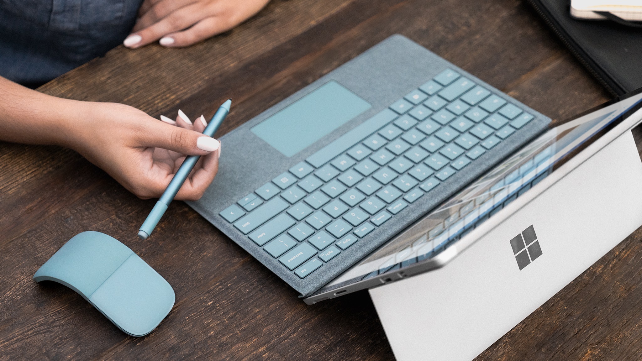 Person sitting at a table holding the new aqua Type Cover, Surface Arc Mouse, and Surface Pen.